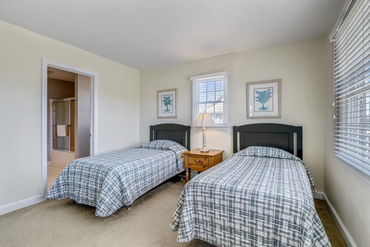 Your bright bedroom features twin beds with crisp plaid linens, a cozy nightstand, and calming coastal artwork that invites peaceful rest after a day of exploring.