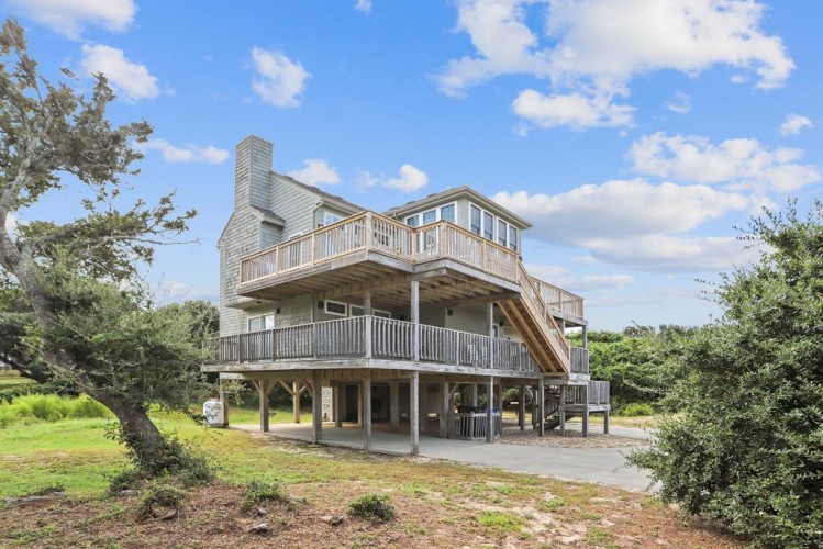 Coastal vacation home elevated on stilts, surrounded by lush greenery and mature trees, offering multiple outdoor deck levels for relaxation and entertainment.