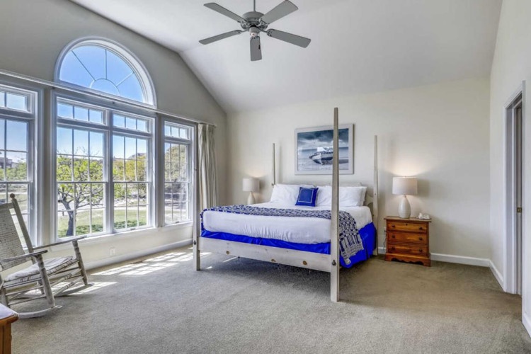 Picture yourself waking to soft morning light streaming through soaring windows. This tranquil bedroom features a plush four-poster bed, soothing coastal artwork, and a gentle breeze from the ceiling fan to lull you into pure relaxation.