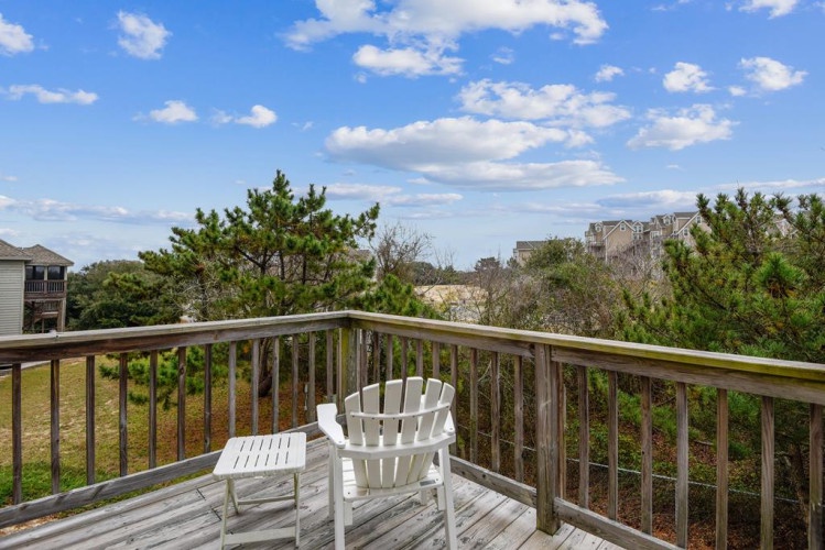 A peaceful coastal setting frames this second-floor deck, where nearby homes nestle among mature pines and native vegetation under open blue skies.