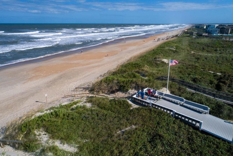 Aerial view of the property's beachfront location, with sandy shores, rolling waves, and easy access to the Atlantic Ocean. A flag marks the pathway leading to the beach.