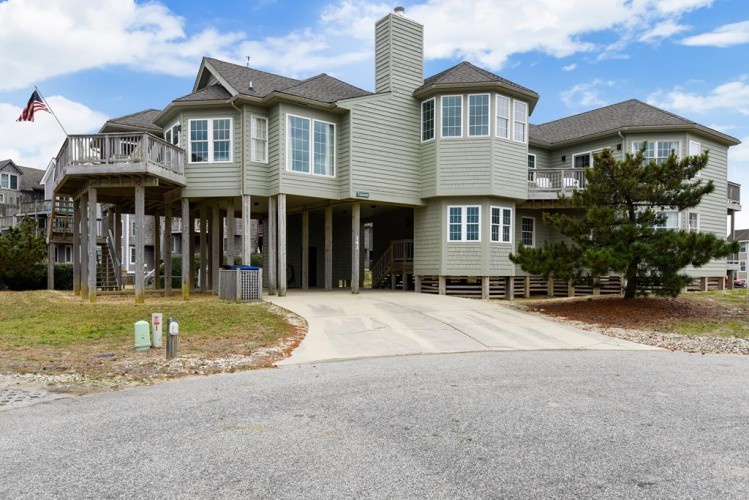 A spacious coastal vacation home elevated on sturdy pilings, featuring wrap-around decks, a turret-style upper level, and ample parking beneath—designed for relaxed beach living in a quiet coastal neighborhood.