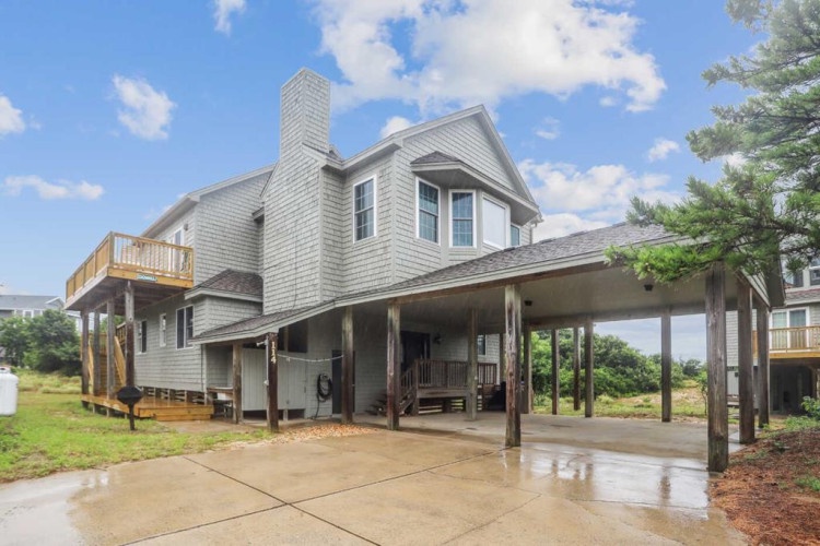 A coastal vacation home features classic shingled siding, an upper-level deck, and covered parking beneath the elevated structure—designed to embrace beachside living in a peaceful, natural setting.