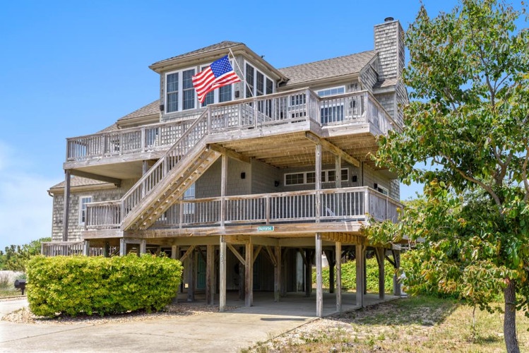 A distinctive coastal property featuring multi-level decks and classic shingled architecture, proudly displaying its beachside character beneath a bright blue sky.