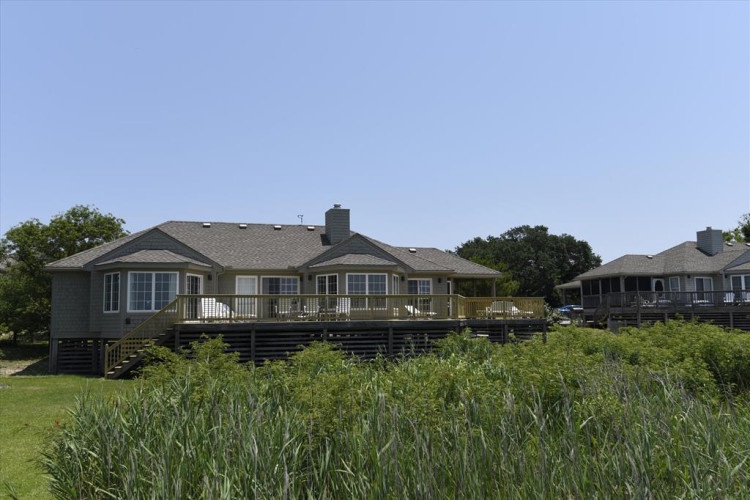 This waterfront vacation home is nestled in a peaceful coastal neighborhood, surrounded by natural grasses and open space. The elevated deck offers expansive views across the serene landscape.