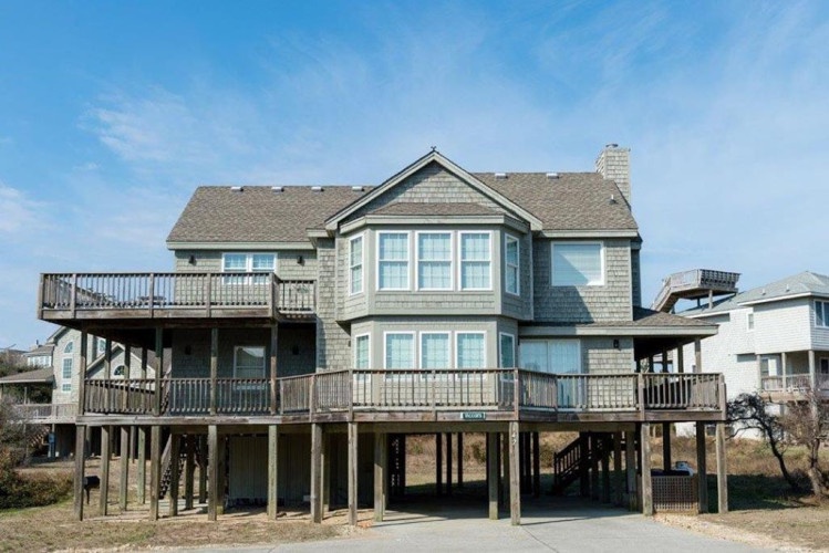 A classic coastal vacation home elevated on stilts with wraparound decks, offering beach access and neighborhood charm under clear blue skies.