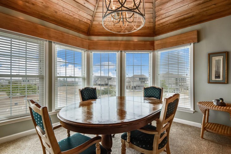 Gather around the elegant wooden dining table under a rustic chandelier, where natural light pours through expansive windows and vaulted wood ceilings create a warm, inviting atmosphere for memorable meals together.