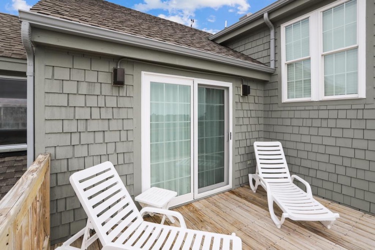 Exterior deck with two lounge chairs positioned beside sliding glass doors against gray shingle siding and blue sky overhead.
