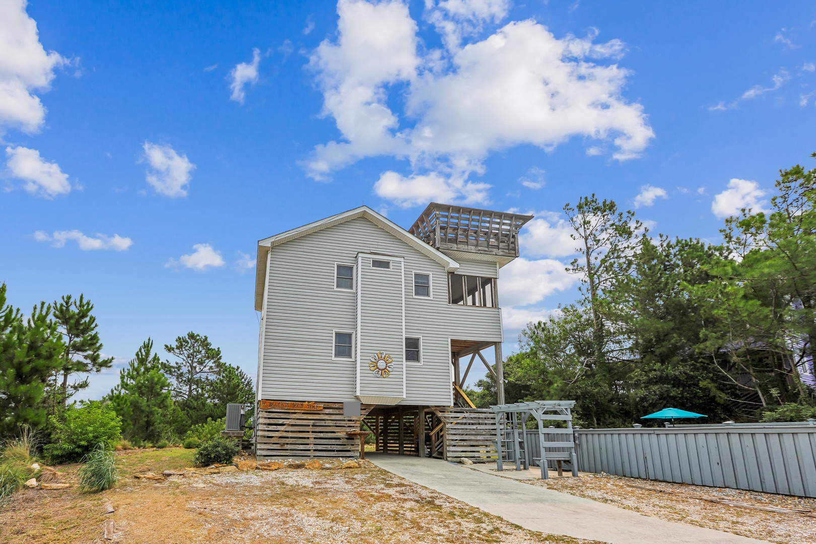 A coastal vacation home with elevated deck and rooftop viewing platform, surrounded by natural greenery and blue skies.