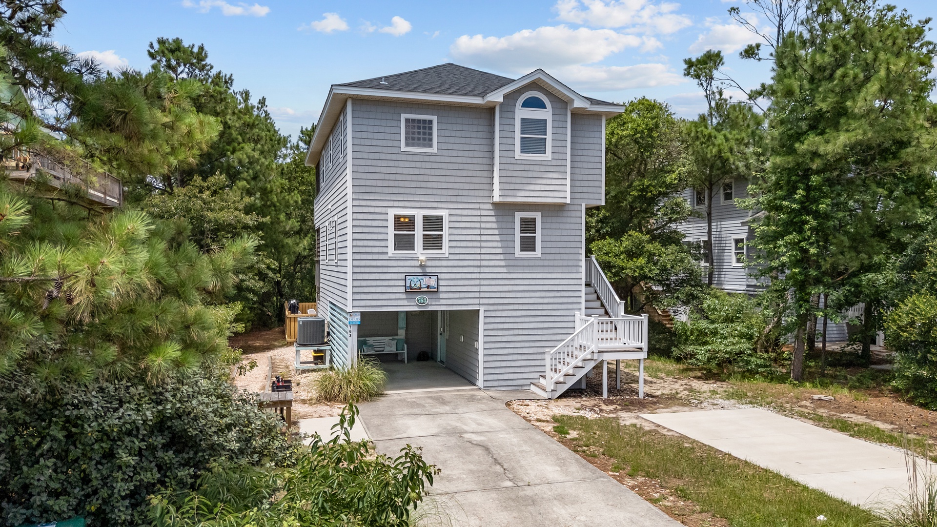 A charming coastal-style vacation home surrounded by lush pine trees, offering convenient covered parking and a welcoming entrance staircase. Located in a peaceful, tree-lined residential area perfect for relaxing getaways.
