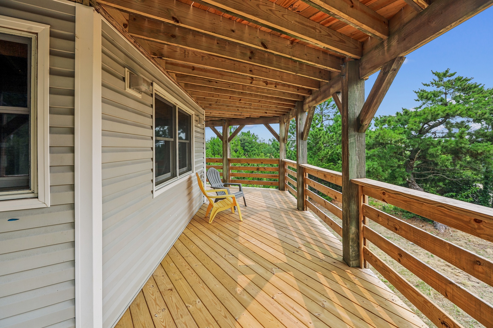 This covered terrace showcases stunning lush greenery through its wooden railing, offering a peaceful outdoor retreat. The exposed beam ceiling and sunny yellow chair invite you to relax and soak in the natural surroundings.