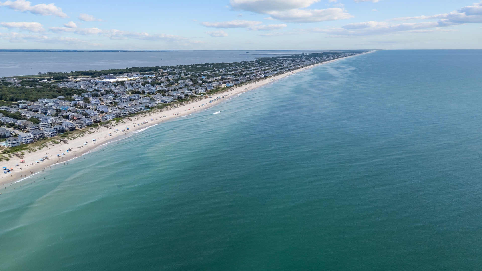 Stunning aerial view captures a pristine beachfront community nestled between turquoise waters and tranquil bay, framed by soft clouds and sunlit shoreline.