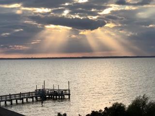 Dramatic sunbeams pierce through clouds over the calm water, with a wooden dock stretching into the peaceful bay—a serene waterfront setting perfect for unwinding.