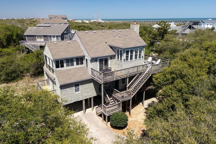 Aerial view of a spacious coastal vacation home nestled among lush greenery, featuring multiple decks and balconies, with ocean views and beach access nearby.