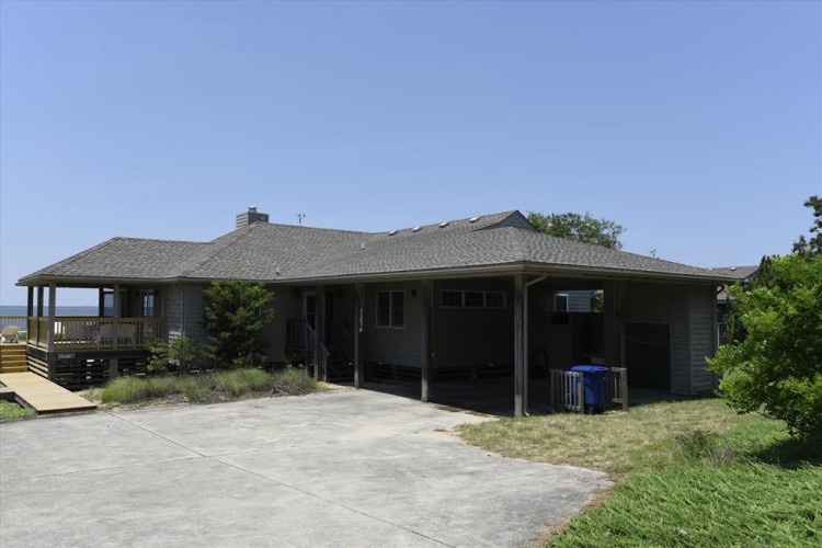 A well-maintained vacation rental featuring a spacious carport and wraparound deck, set in a coastal area with greenery and ocean access nearby.