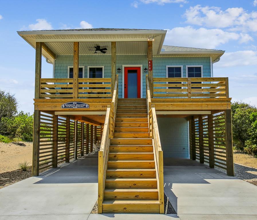 Charming beachside cottage elevated on stilts with coastal blue siding, a welcoming red door, and spacious front deck overlooking natural dunes and coastal vegetation.