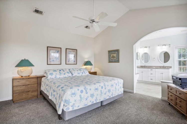 Your serene bedroom features vaulted ceilings with a ceiling fan, a king-sized bed dressed in coastal linens, and classic bedside lamps. An ensuite bathroom awaits through the archway, combining comfort and coastal charm.