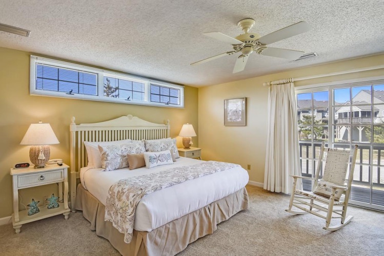 Sink into crisp linens and wake to gentle sunlight streaming through coastal windows—your serene retreat awaits with charming rocking chair and soft coastal décor.