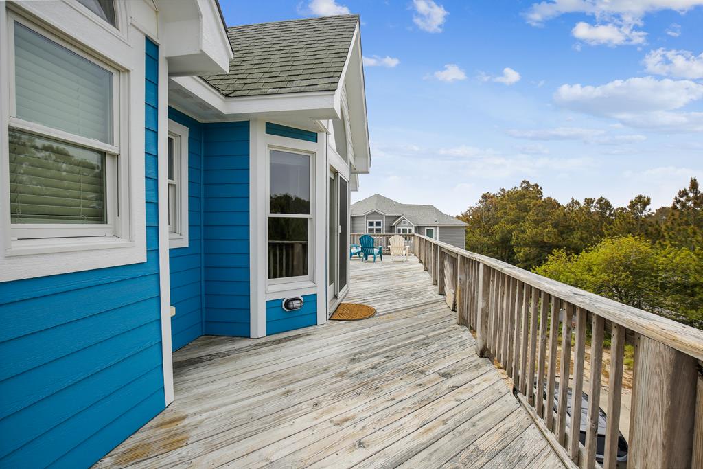 Bright blue coastal exterior with a wraparound deck featuring weathered wood, neighboring homes, and lush greenery under a sunny sky—ideal arrival setting for a beach getaway.