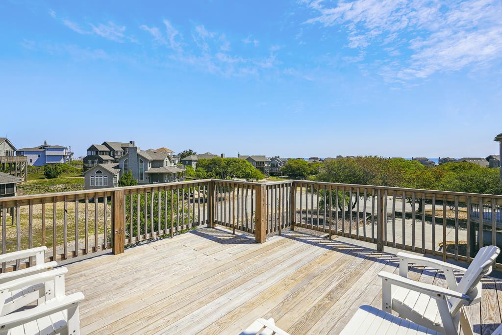 A spacious rooftop deck with wooden railings offers expansive views of the coastal neighborhood and nearby beach homes. The outdoor space features lounge chairs for relaxation under open skies.