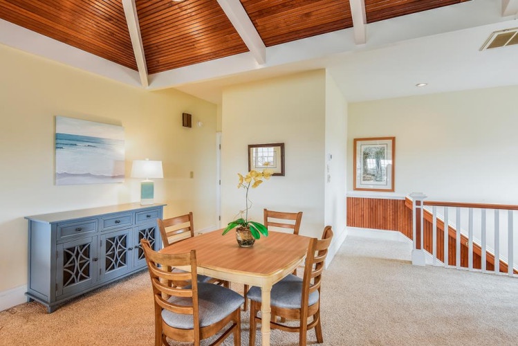 Gather at the rustic wooden dining table under soaring wood-paneled ceilings, where warm natural light and beachy artwork create a relaxed coastal retreat.