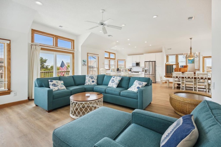 Spacious open-plan living with vaulted ceilings, teal sectionals, and coastal décor invites relaxation. Bright natural light flows through large windows, seamlessly connecting the dining area and kitchen for effortless entertaining.
