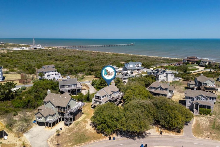 Aerial view of a coastal neighborhood featuring beachfront vacation rentals, natural dune vegetation, and a nearby fishing pier extending into calm blue waters.