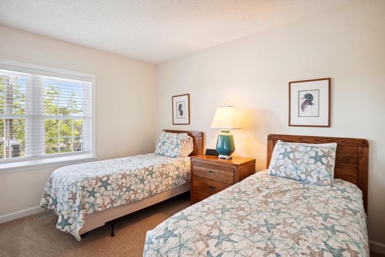 Relax in your light-filled bedroom featuring two cozy twin beds dressed in coastal-themed linens, warm wood furnishings, and charming artwork that welcomes you to unwind after a day exploring.