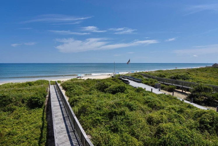 Elevated boardwalks wind through coastal dunes toward a pristine sandy beach and calm turquoise waters under clear blue skies.