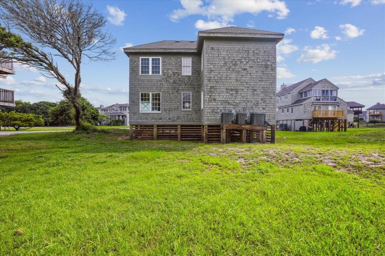 This coastal vacation rental features weathered gray shingle siding, elevated on stilts with a spacious lawn and mature trees, nestled in a quiet neighborhood near other beach homes.