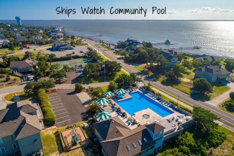 Aerial view of the property situated along the coastal road, with its community pool and parking area, neighboring a quiet residential area near the waterfront.