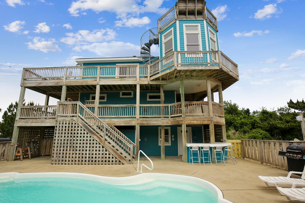 This distinctive turquoise beach house features expansive multi-level decks, a private pool, BBQ area, and rooftop observation tower overlooking coastal vegetation.