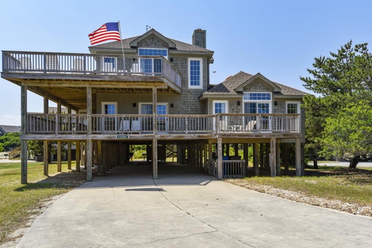 Coastal vacation rental elevated on sturdy pilings with spacious covered parking, wraparound decks, and an American flag welcoming visitors to this beach retreat.