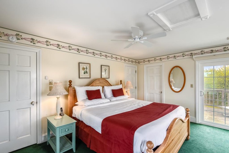 Step into your serene retreat with a welcoming wooden bed dressed in crisp linens and rich red accents, soft carpeting beneath your feet, and natural light pouring through glass doors.