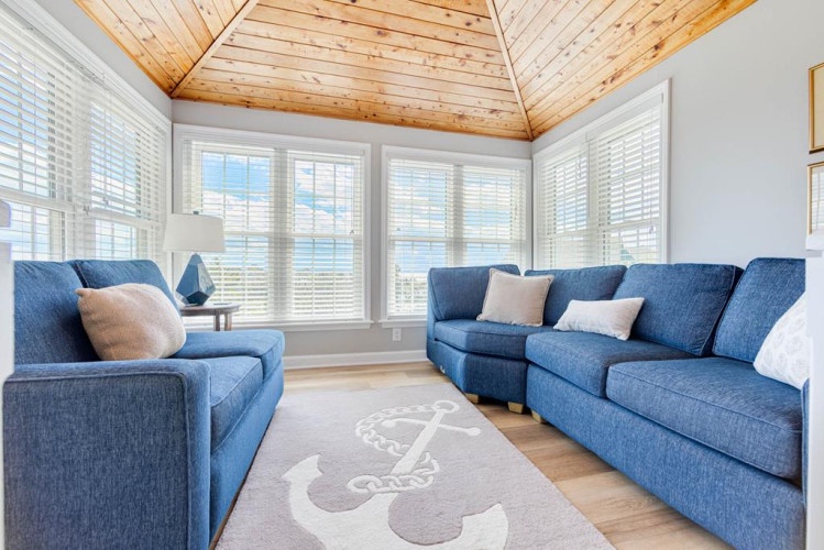 Sink into plush blue sofas beneath warm wood ceilings in your private living room, where bright windows frame serene views and coastal charm surrounds you.