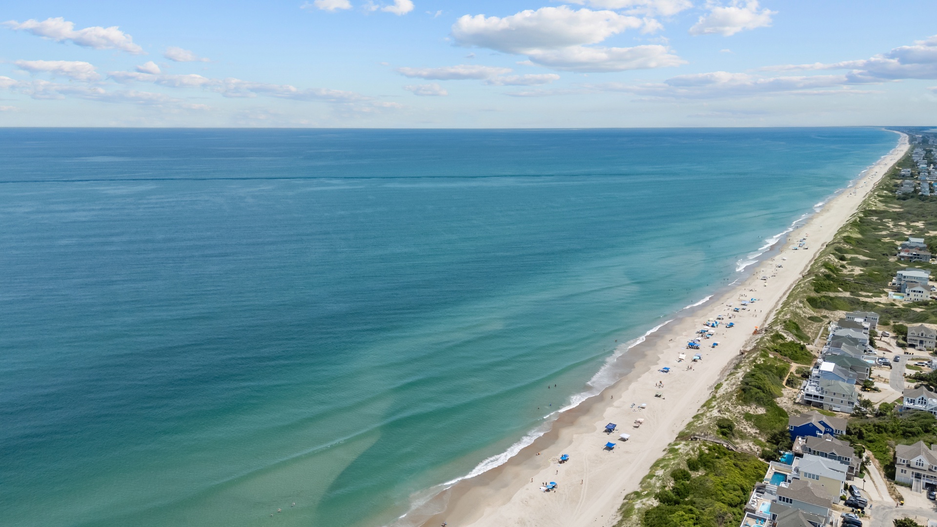 Stunning aerial perspective showcases pristine coastal beauty with turquoise waters meeting white sand beach, dotted with umbrellas and bordered by charming beachfront homes along the shoreline.