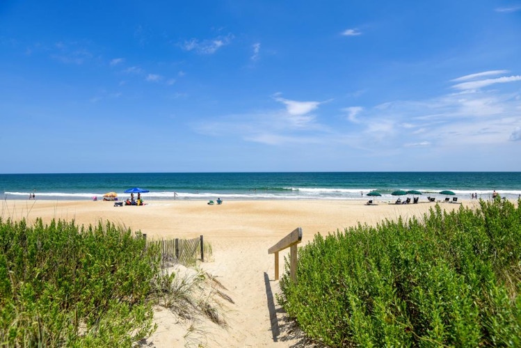 Sandy path leads through natural dunes to a wide, pristine beach with gentle ocean waves under a clear blue sky—an idyllic coastal escape just steps away.