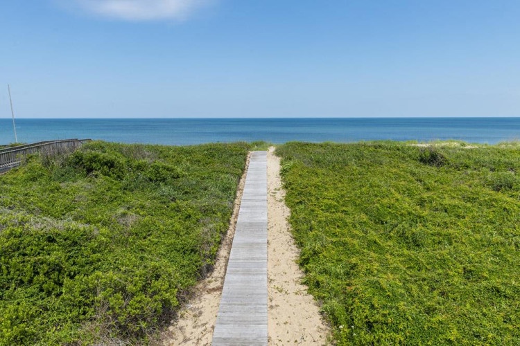 A wooden boardwalk leads through coastal dunes directly to the sparkling sea, framing an idyllic beachfront location surrounded by natural greenery.