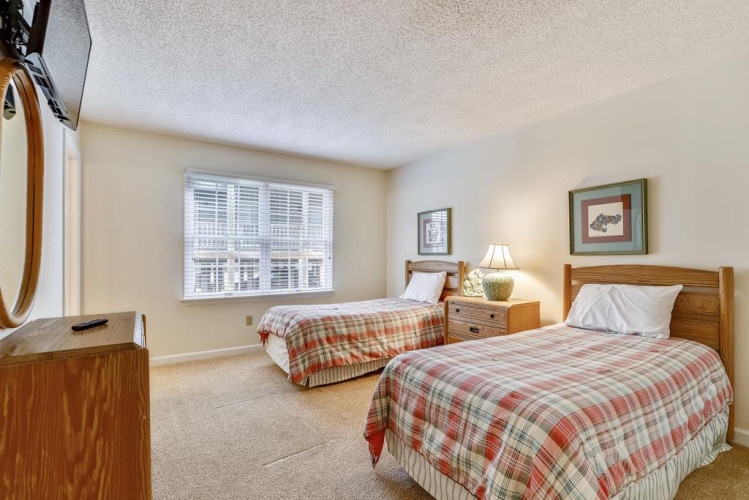 Your cozy bedroom features two plush twin beds with warm plaid linens, natural light from large windows, and rustic wood furnishings that create a relaxing mountain retreat atmosphere.