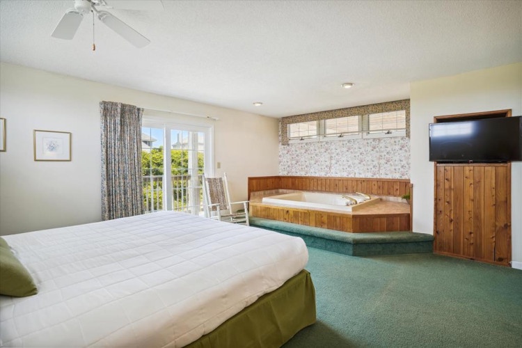 Your spacious bedroom features a plush bed, private hot tub for ultimate relaxation, ceiling fan for comfort, and balcony access with natural light streaming through elegant curtains.
