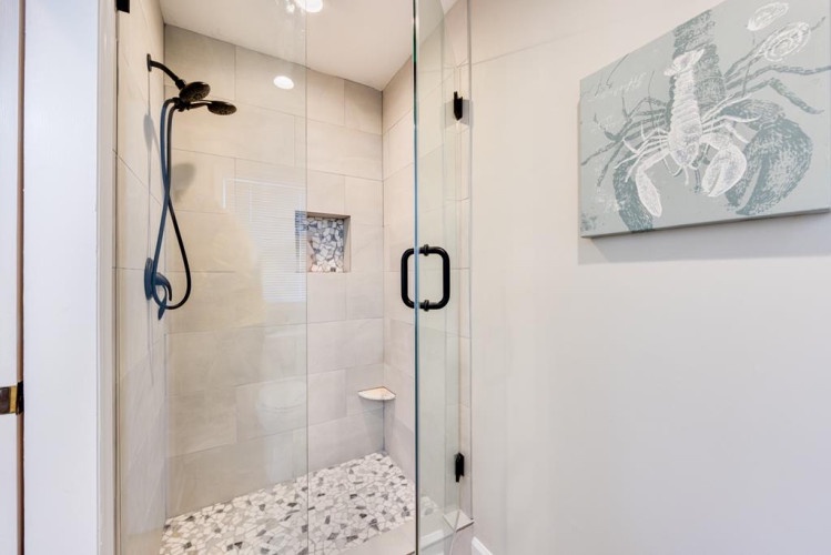 Step into your private retreat—a sleek walk-in shower with a rainfall head, pebble flooring, and coastal-inspired artwork that invites relaxation after a day by the sea.