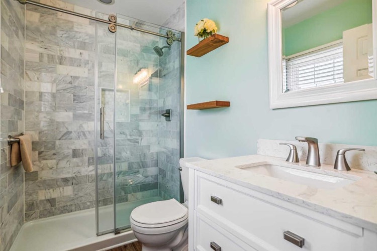 Your private bathroom features a sleek walk-in shower with marble-look tile, modern fixtures, and soothing mint walls. Fresh towels and floating shelves add convenience and charm.