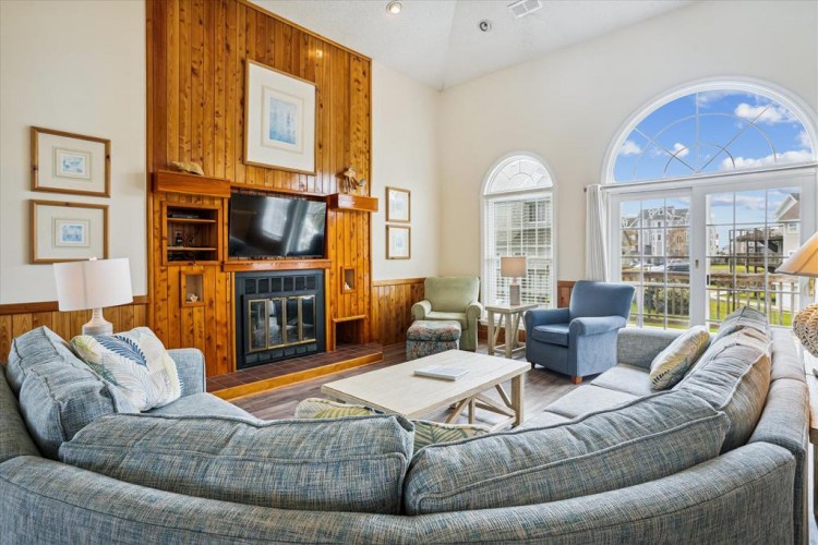 Unwind in your spacious living room, where coastal charm meets comfort with plaid sofas, a cozy fireplace, and arched windows framing beautiful neighborhood views.