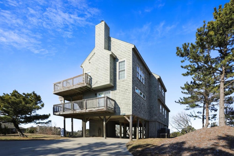 Coastal vacation home elevated on sturdy pilings with wraparound decks offering elevated views of the surrounding landscape and blue skies.