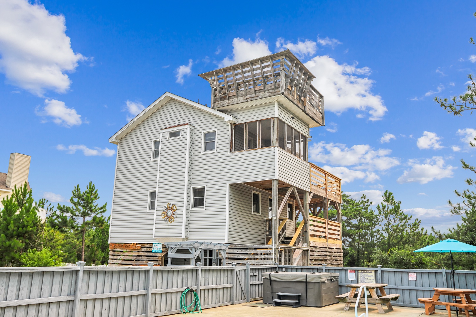The property features a distinctive multi-story structure with private upper-level decks and an observation tower, set among coastal pines with a ground-level patio space including hot tub and outdoor seating.