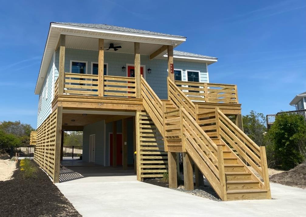 Modern beach house on stilts with covered parking, expansive deck, and coastal-style architecture situated near natural vegetation.