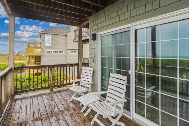 A charming covered deck awaits with rocking chairs overlooking open fields and neighboring homes under bright blue skies—perfect for relaxing after exploring the area.