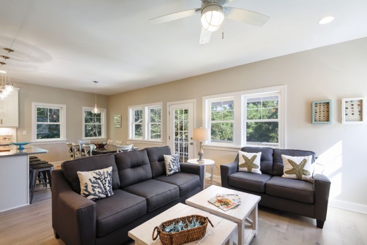 Step into your sunlit retreat where sleek charcoal sofas invite relaxation. Large windows flood the space with natural light, while the ceiling fan keeps you cool as you unwind in this modern, airy haven.