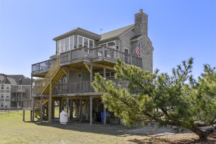 Coastal vacation home elevated on stilts with wraparound decks and an American flag, set among maritime pines and neighboring beach residences.
