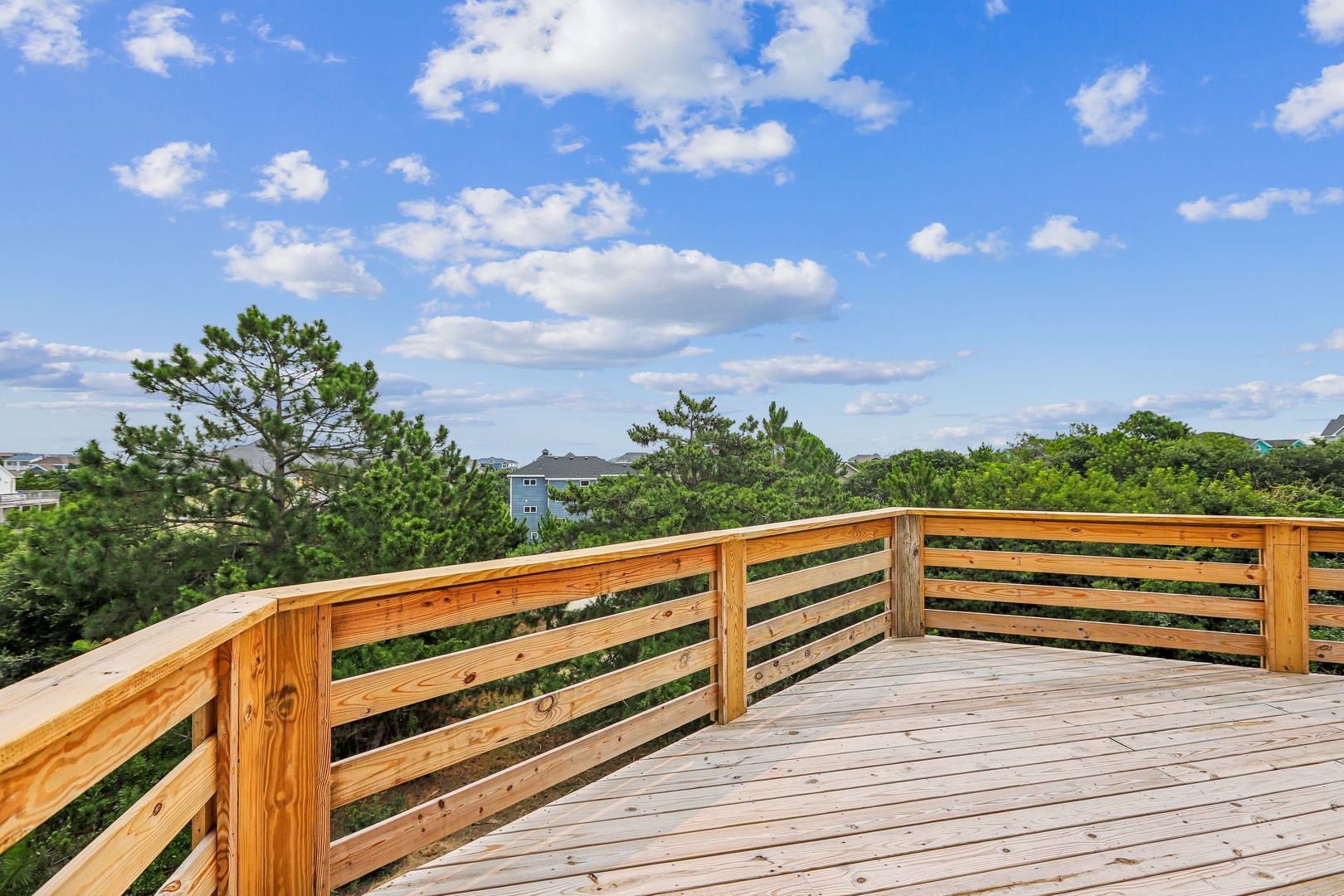 A spacious wooden deck overlooks lush greenery beneath a bright, clear sky. The open railing design frames sweeping views of trees and neighboring homes, creating your perfect outdoor retreat.